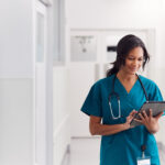 Female Doctor Wearing Scrubs In Hospital Corridor Using Digital Tablet